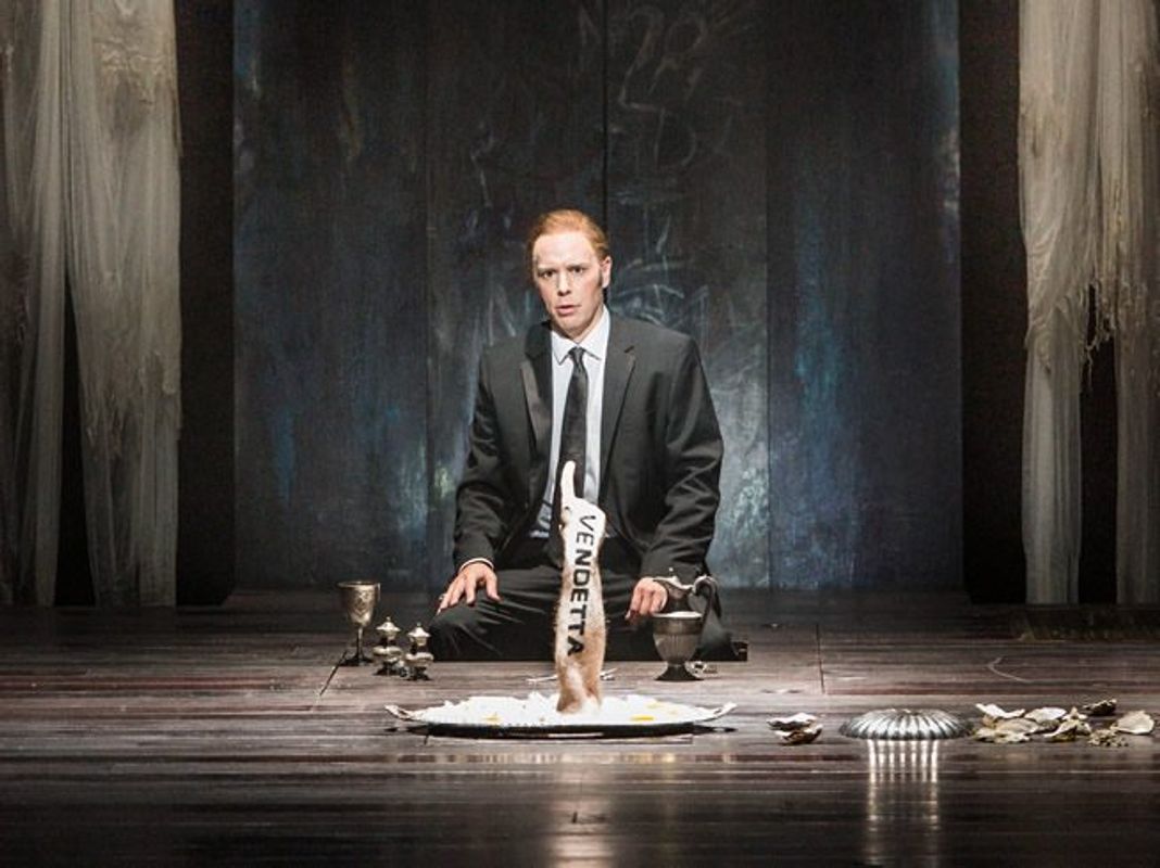 A person is sat on the floor infront of a silver tray. There is an arm coming out of the tray and it has the work 'Vendetta' written on it.