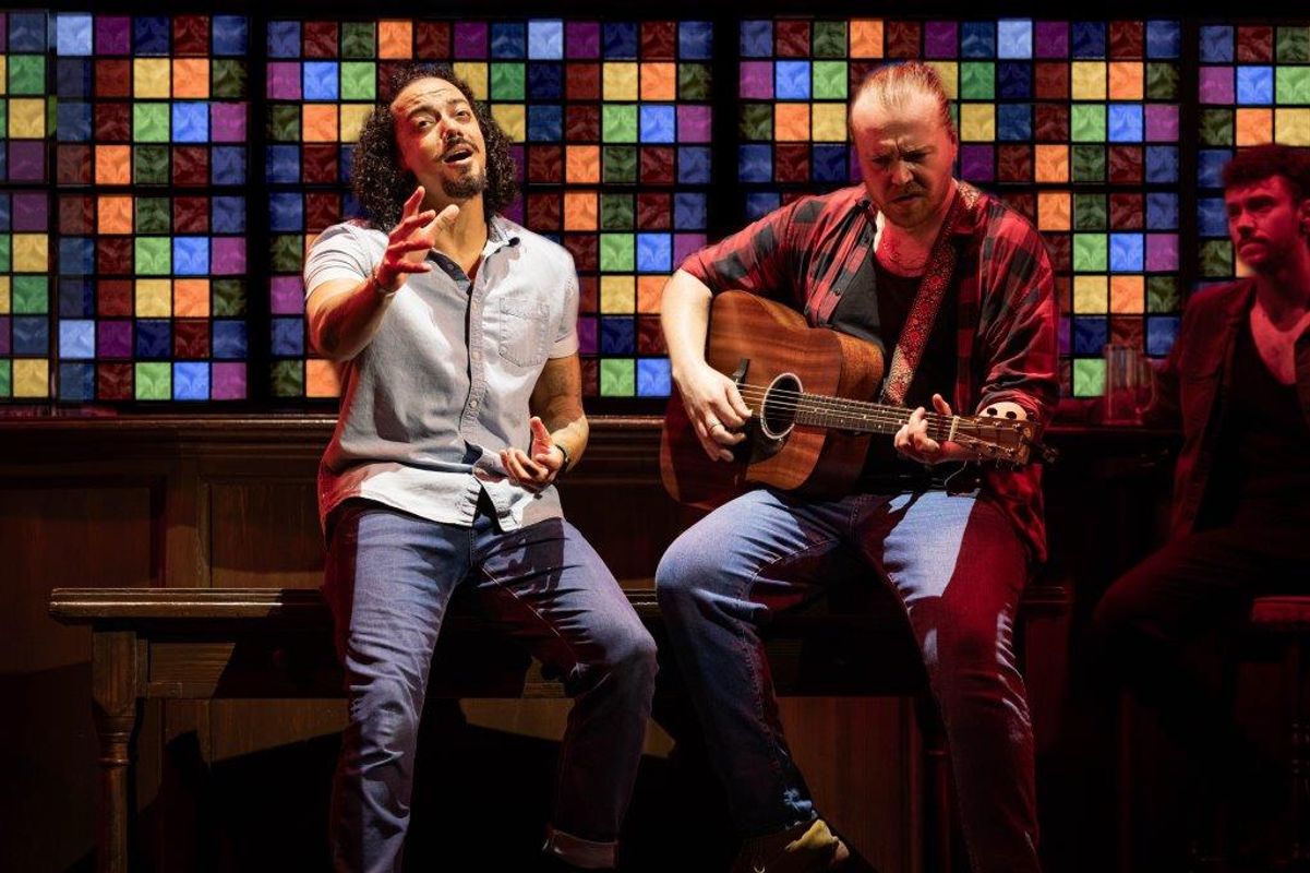 Three people sat at a bar, one is singing and the other playing a guitar