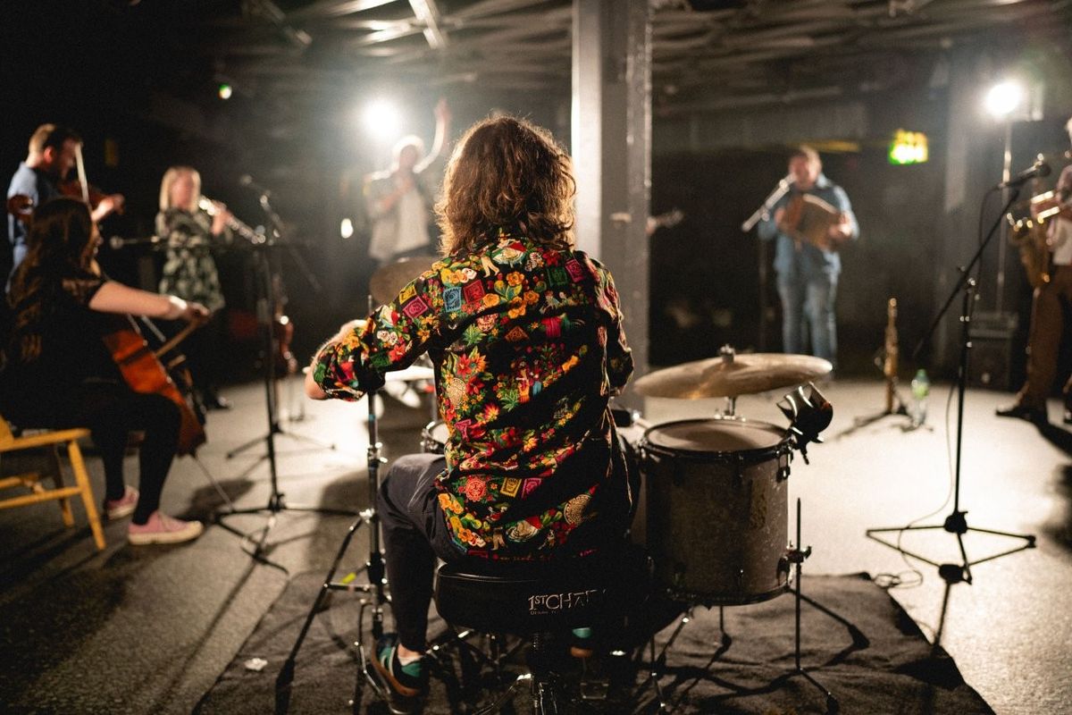A person with curly hair, wearing a colorful floral shirt, plays drums on stage. Several musicians are setting up or tuning their instruments, including a cello and brass instruments. The setting appears to be a dimly-lit music venue with stage lights shining on the performers.