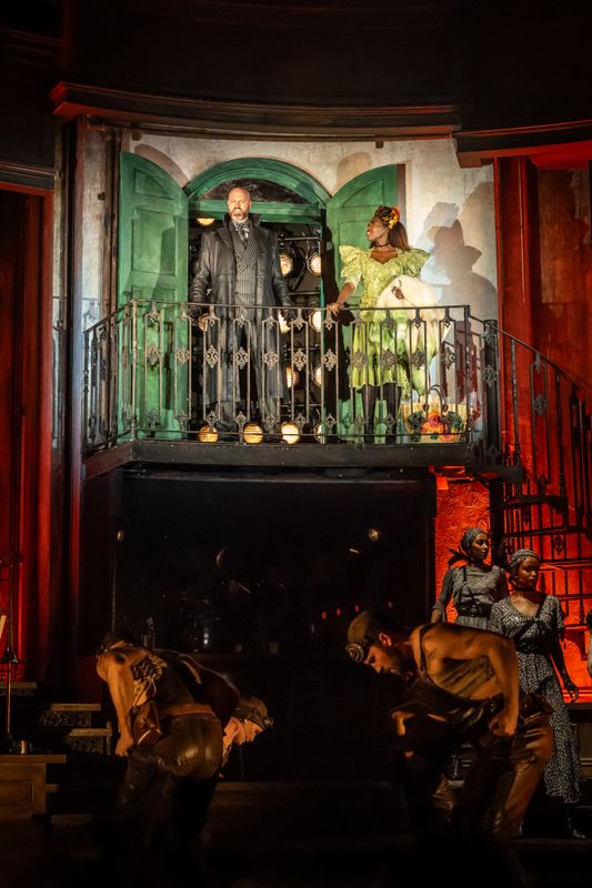 Two actors, one wearing a leather blazer and the other wearing a green dress, stand on a balcony  overlooking ensemble members.