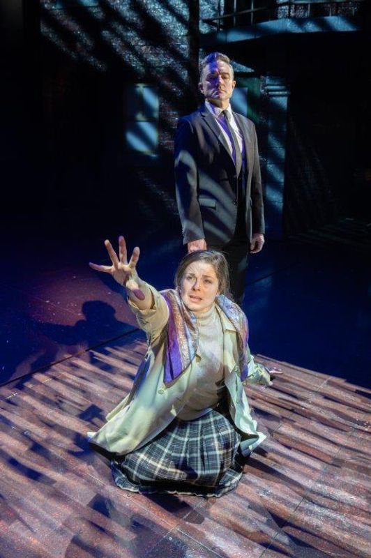 There is a woman kneeling on the floor with a hand up in the air. A man in a suit is stood behind her