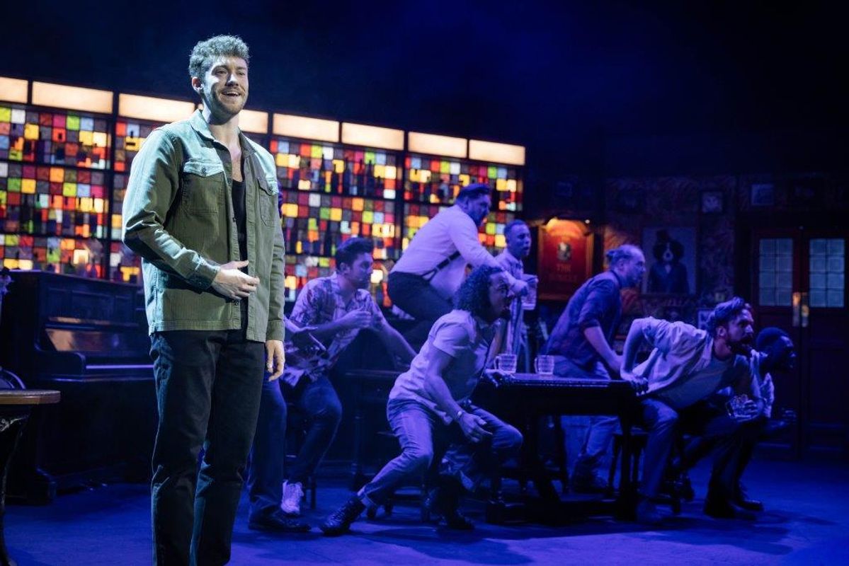 Person stood to the side with a spot light on them, there is a group in the background sat round a table