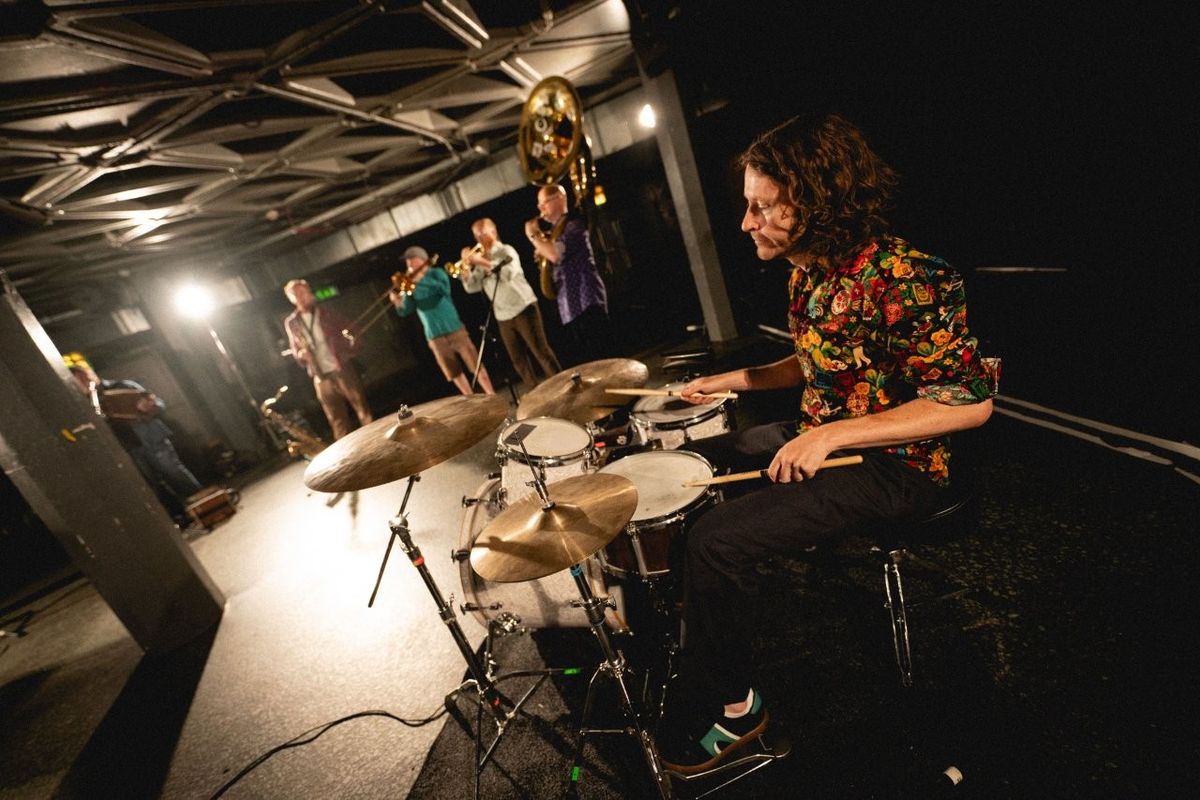 A person playing a drum set in a dimly lit room, with a group of people in the background playing various brass instruments.