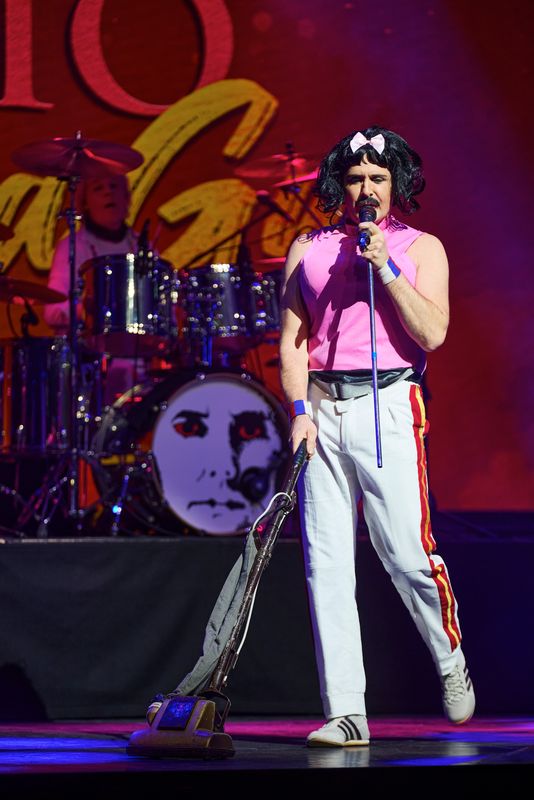 A man singing on stage and wearing a pink vest top, with a black wig