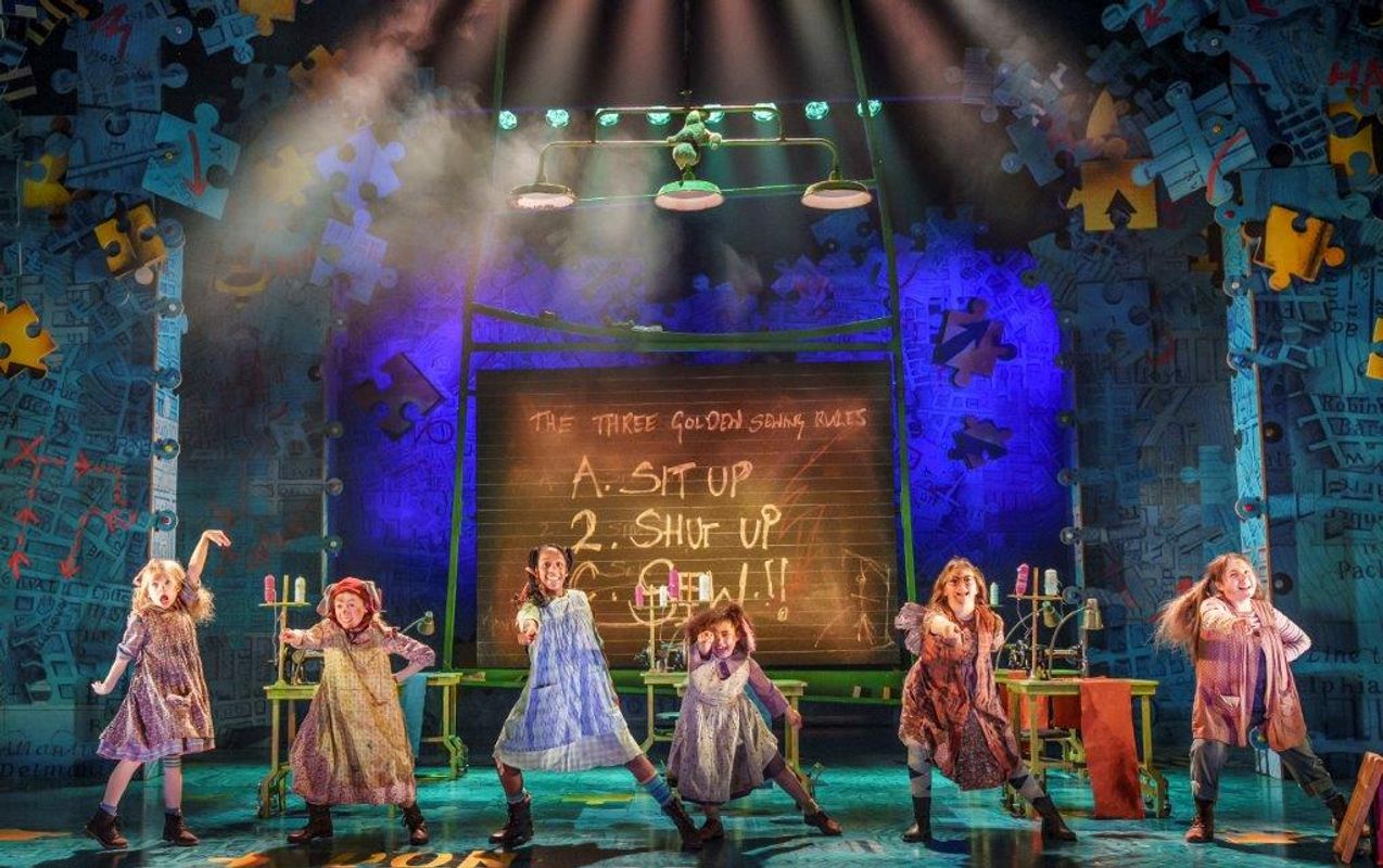 Theater scene from "Matilda: The Musical" with six children in ragged costumes dancing energetically on a colorful, puzzle-themed stage.
