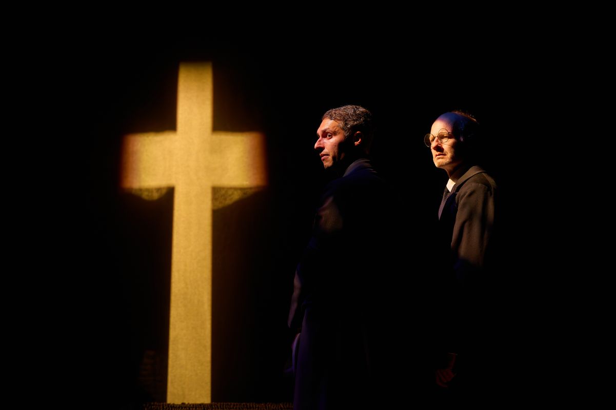 Two people dressed in black stand next to a cross.