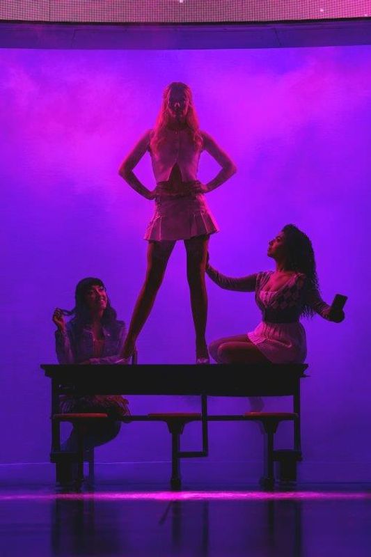 Three women pose dramatically on a stage with vibrant purple lighting. One stands confidently on a table, hands on hips, while the other two sit beside her.
