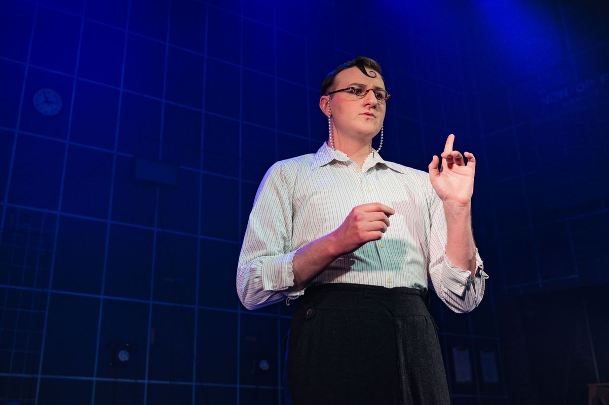 A person stands on stage under blue lighting, wearing glasses and a white shirt, with hands raised expressively. A clock and clipboards are visible on the grid-patterned wall behind, suggesting a dramatic or theatrical setting.