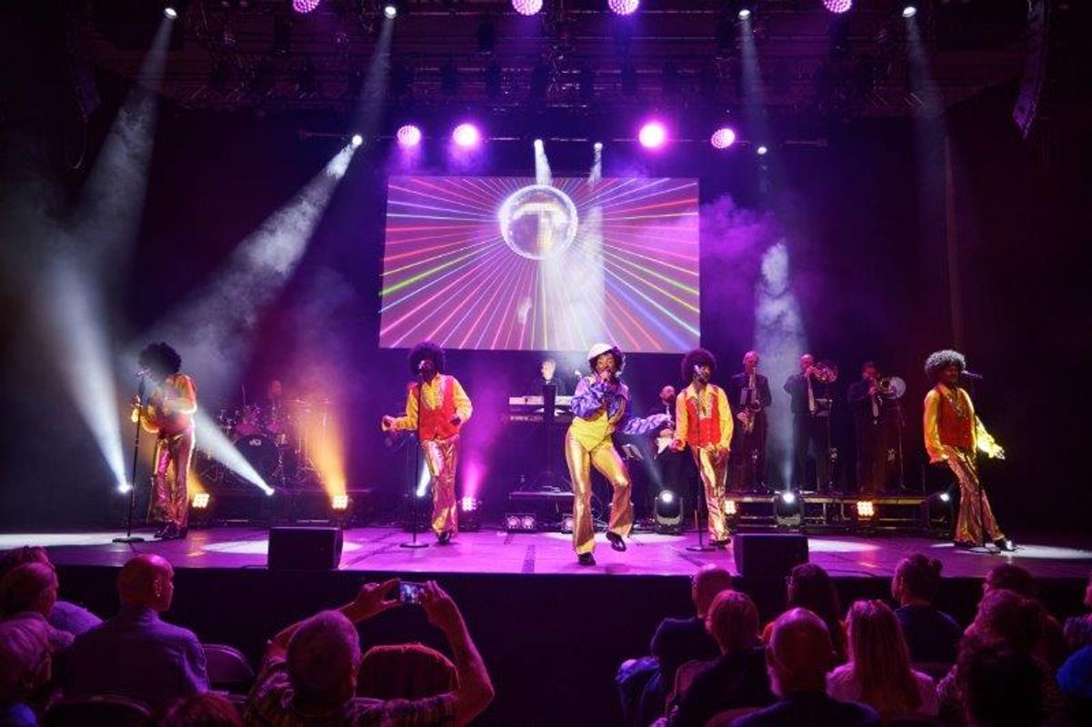 Five singers, in flared trousers, are stood on stage singing.