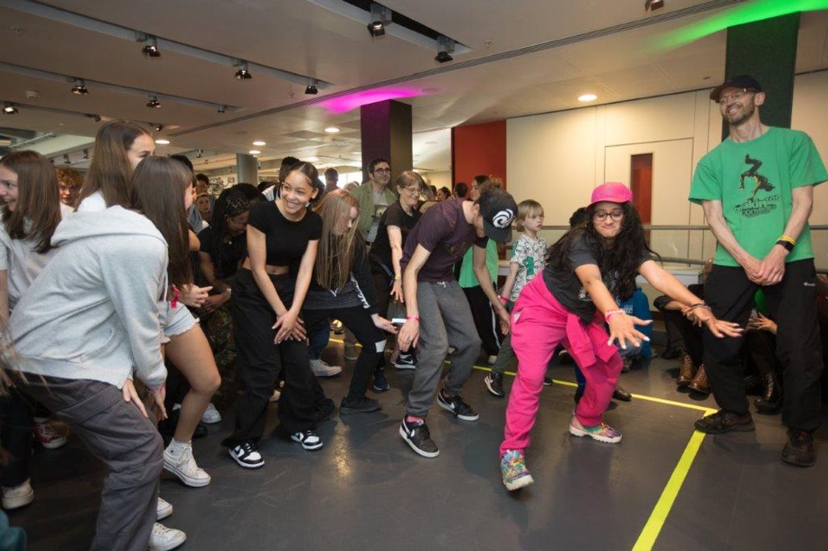 A person wearing a pink hat dances whilst a group of young people stand behind copying the moves.