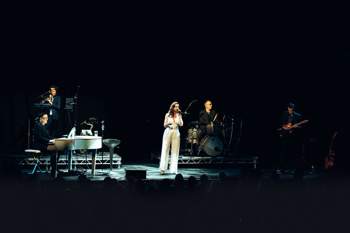 A person wearing a white jumpsuit stands centre stage singing.