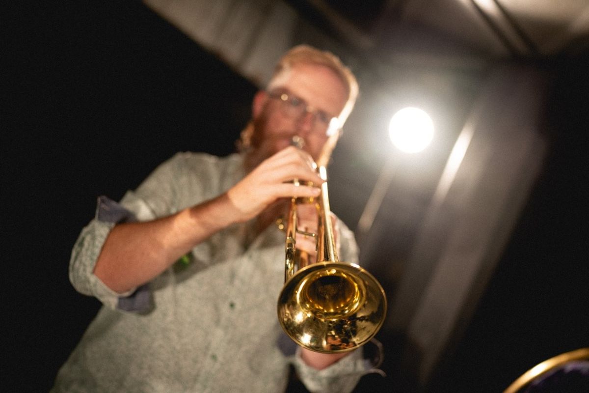 A person is playing a trumpet under stage lighting, with a bright spotlight behind them. The trumpet gleams in the light. The background is dark, highlighting the musician and the instrument.