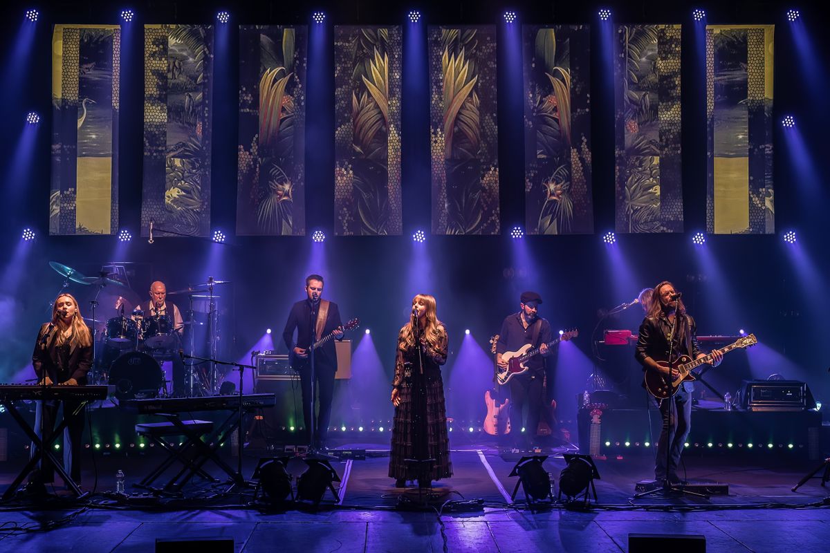 Seven people play instruments on stage with floral images on screens above.