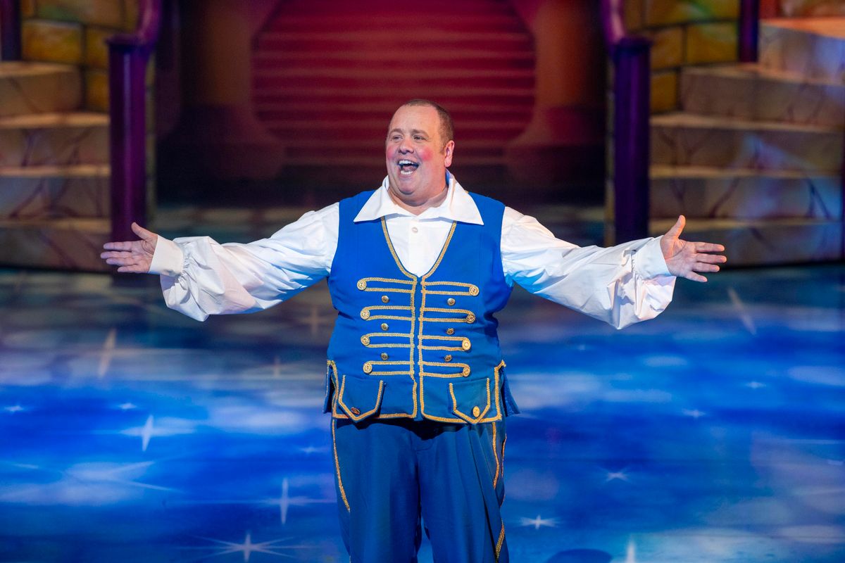 Buttons stands with his arms out to the side wearing a white collared shirt with a blue waistcoat.