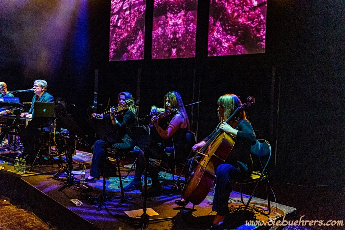 A band performs on an atmospheric stage with purple abstract visuals projected behind them. Musicians play keyboards, violin, cello, and more.