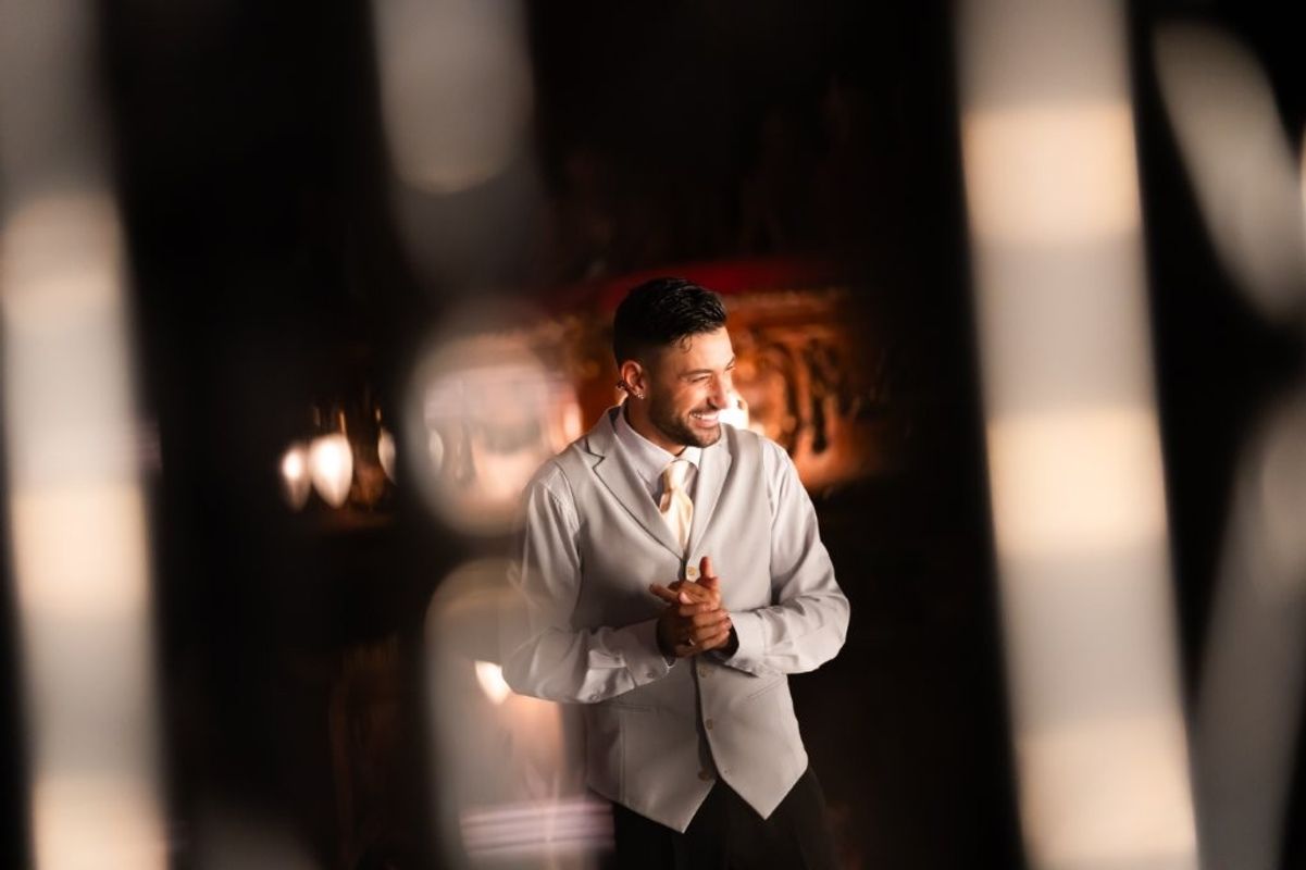 Giovanni wearing a white suit, he is holding his hands Infront of him and smiling away from the camera