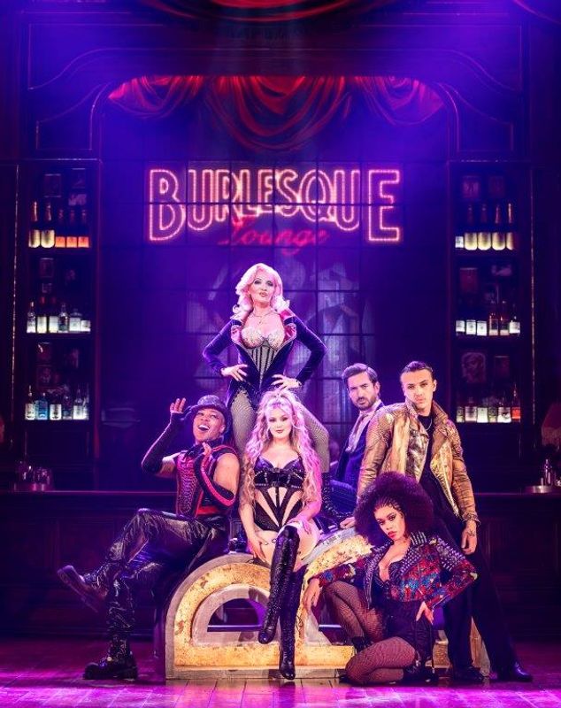A lady is center stage wearing a corset and coattail jacket. Others are sat around her in vibrant and edgy outfits