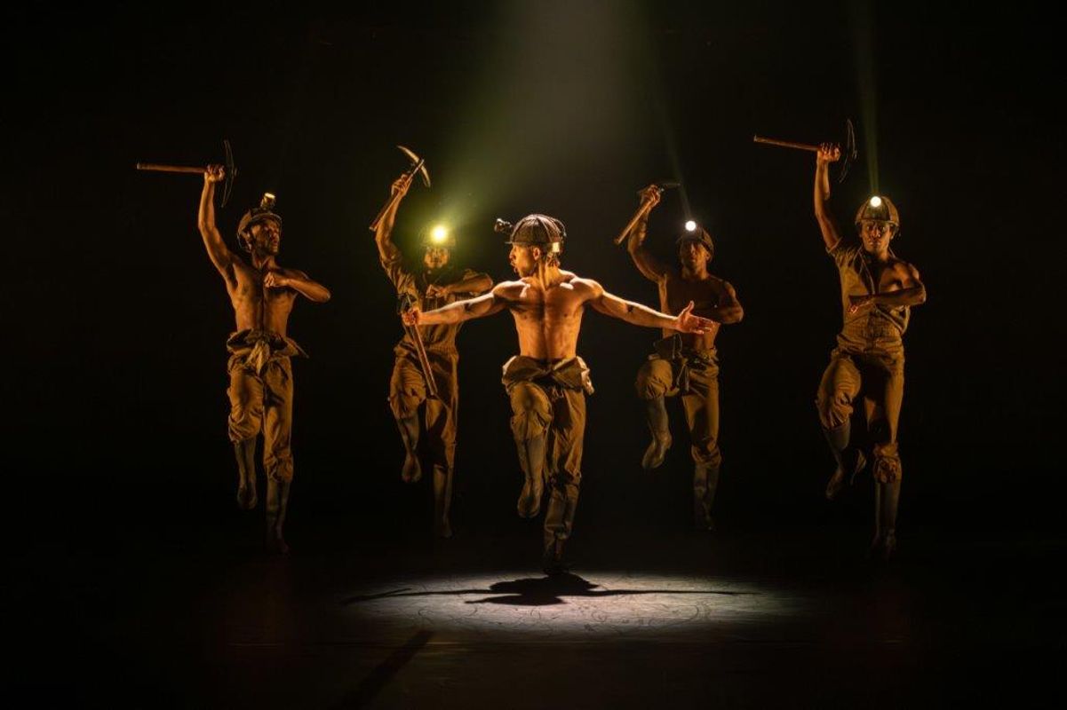 Group of dancers on stage wearing hard hats and holding pick axes above their heads
