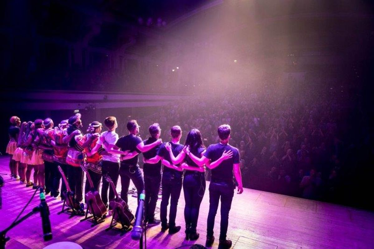 All the performers are stood together in a line with their arms around each other facing the audience