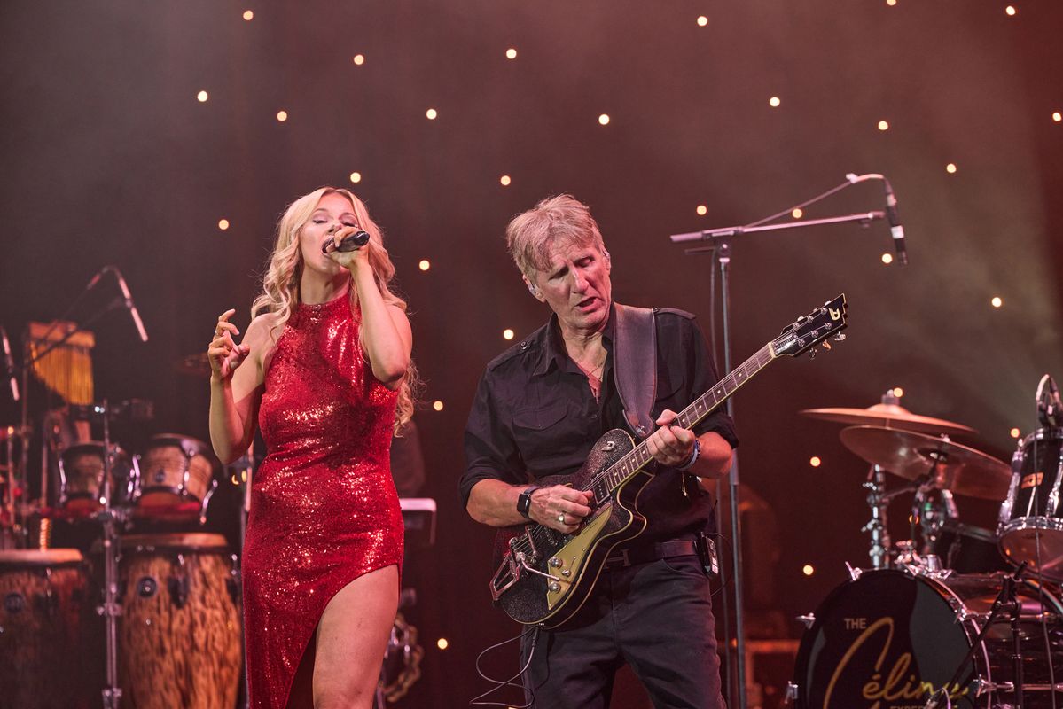 A person in a red sequined dress sings into a microphone. Another person beside them plays an electric guitar. The background features star-like stage lighting. Musical equipment and drums are visible on stage.