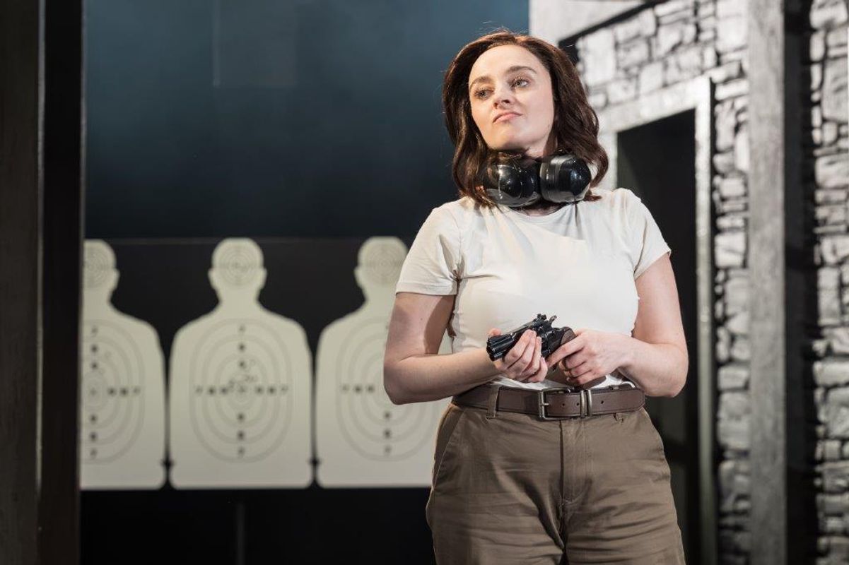A woman is stood in a shooting range. She his holding a gun and has ear defenders around her neck