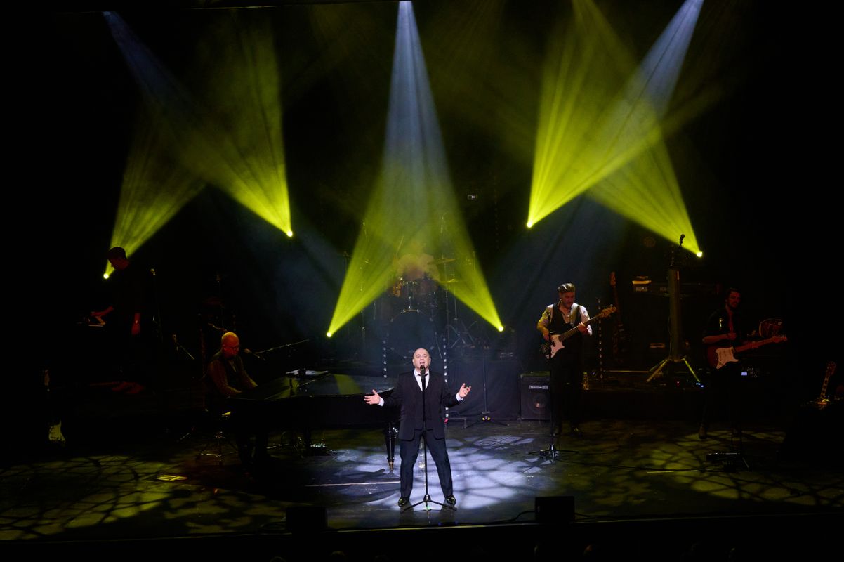 A person in a black suits stands centre stage with yellow spotlights behind.