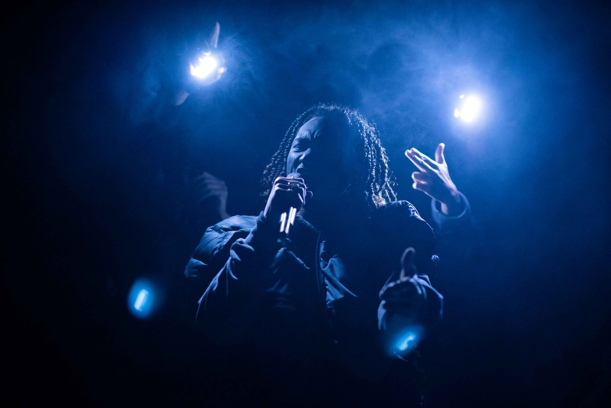 A dark figure lit by two spotlights raises their left hand pointing two fingers.