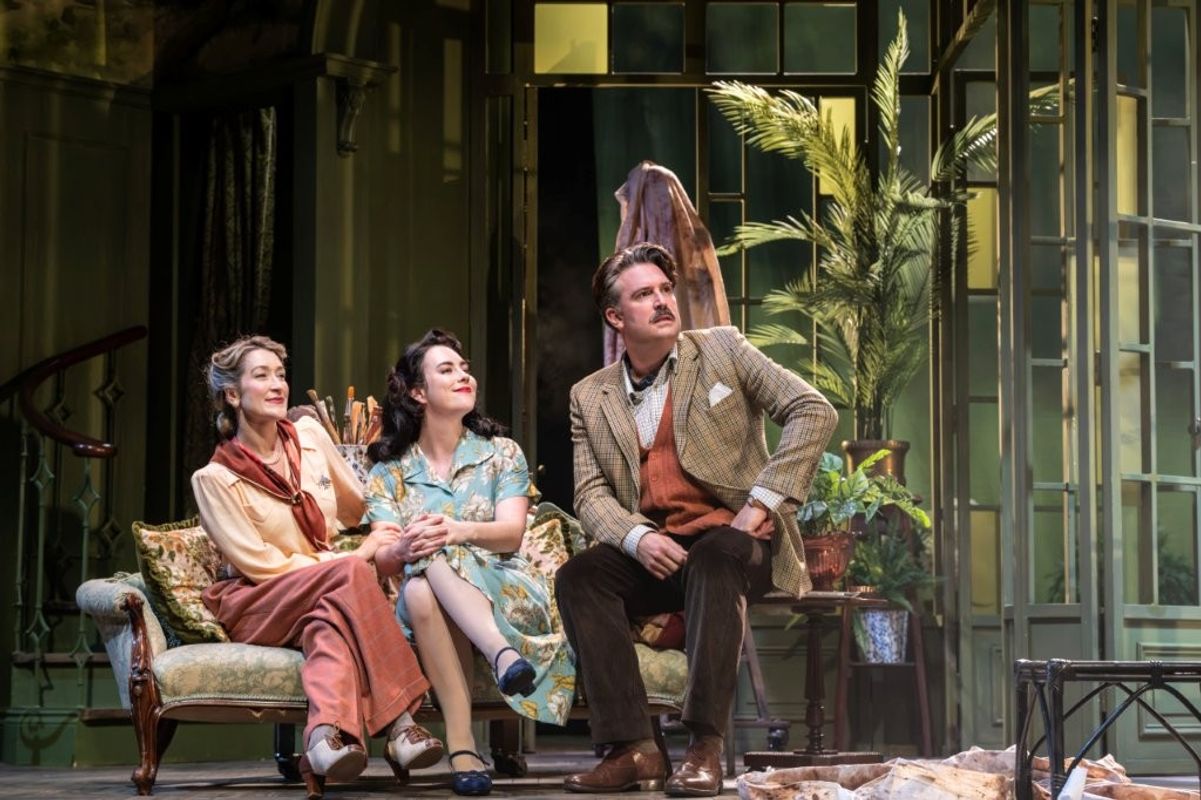 Three people are seated on an elegant vintage sofa in a charming room with large windows and lush plants. They are dressed in period costumes, appearing contemplative and relaxed.