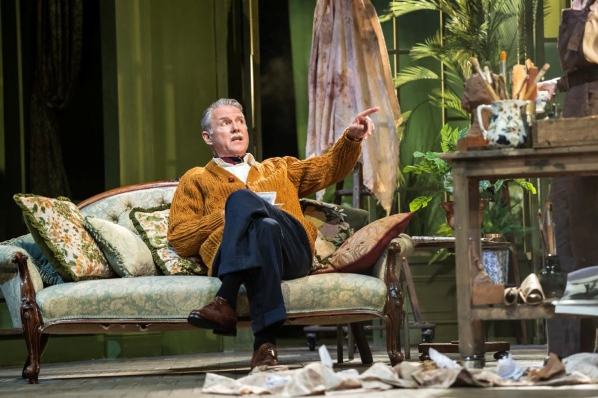 A person wearing a brown cardigan is seated on an elegant sofa on a stage set, pointing towards another person who is partially visible. The setting includes ornate cushions, potted plants, and a wooden table cluttered with art supplies.