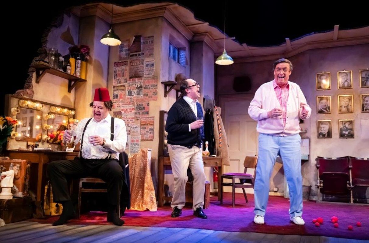 Three actors perform in a lively theater scene. One sits wearing a fez, another stands mid-laugh with glasses, and the third smiles in pastel clothing. The room is adorned with posters and framed photos. The setting is warmly lit, creating a comedic and vibrant atmosphere.