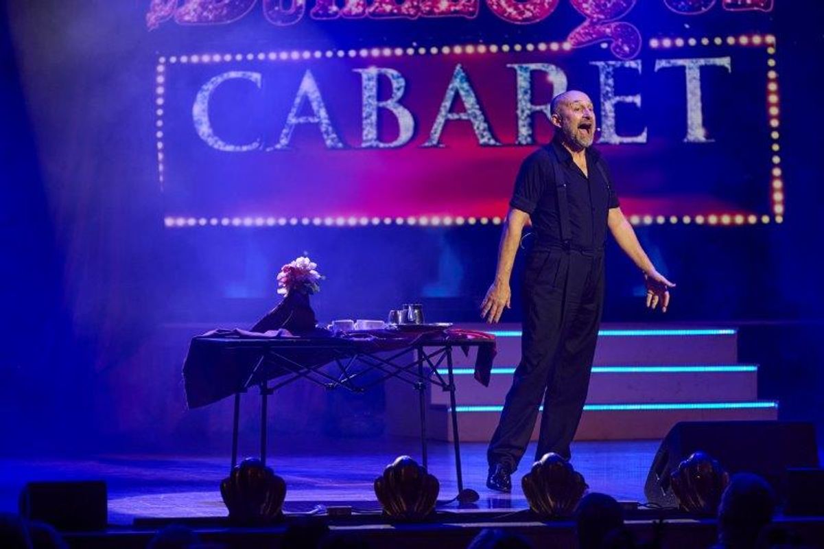 A person in a black shirt and braces stands next to a table.