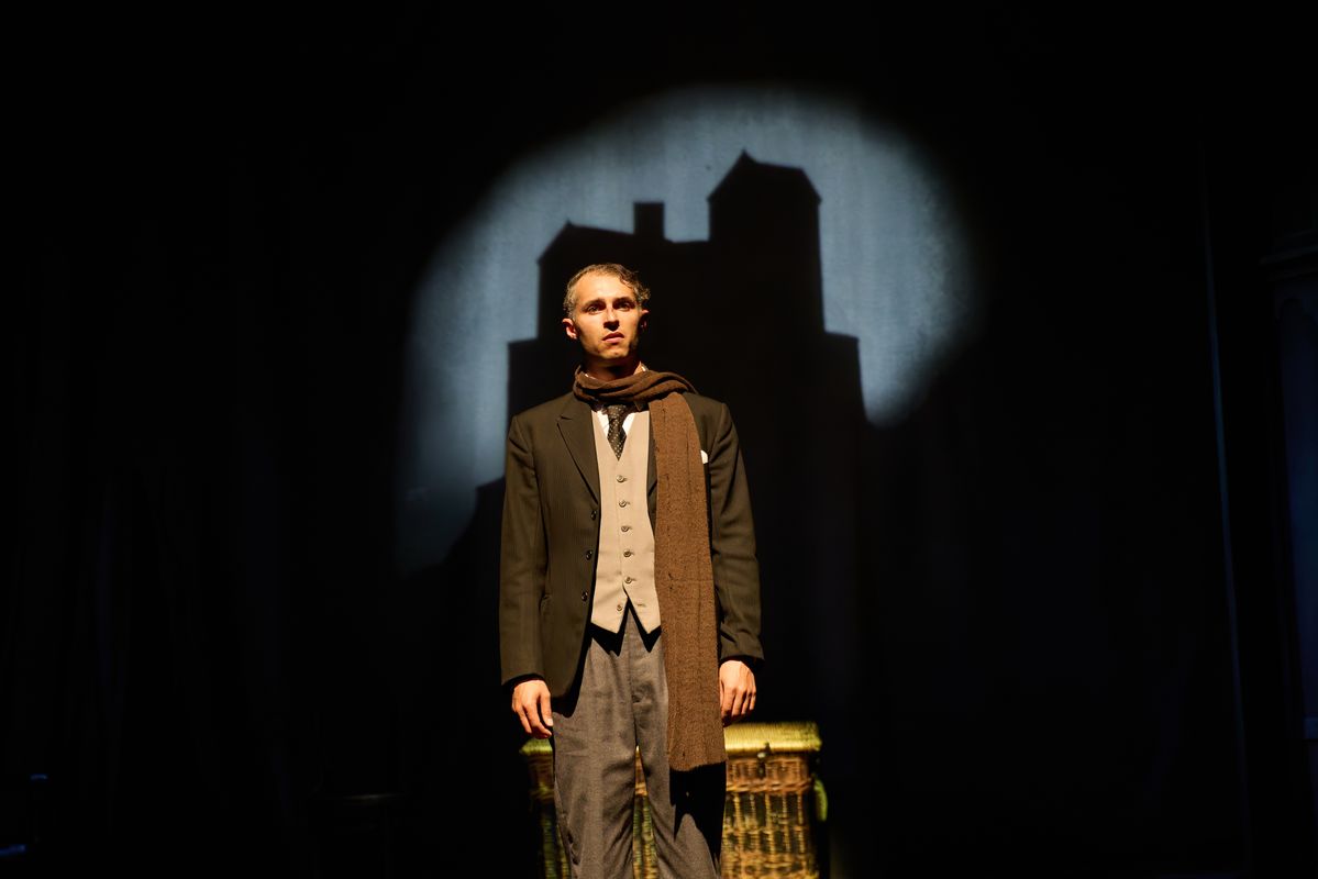 A person wearing a black blazer and brown scarf stands with a house projected in the background.