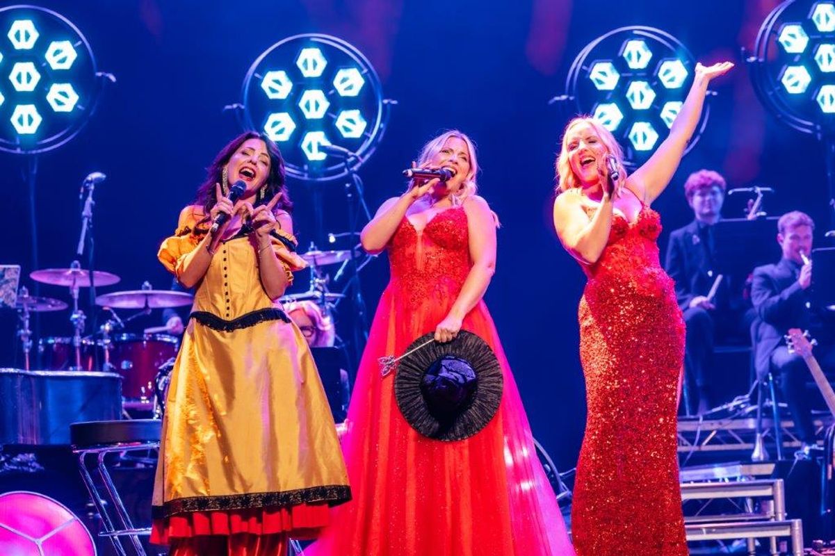 Kerry Ellis, Louise Dearman and Rachel Tucker perform energetically on stage, each holding a microphone. They wear vibrant gowns—two in red and one in yellow. Bright stage lights illuminate the scene, creating a lively and celebratory atmosphere.