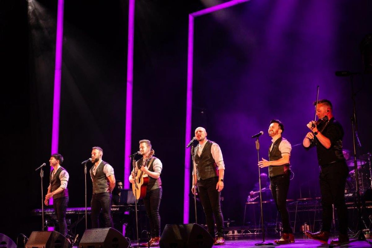 Five male performers in matching vests and shirts sing and play instruments onstage. Purple lights and smoky atmosphere create an energetic concert vibe.