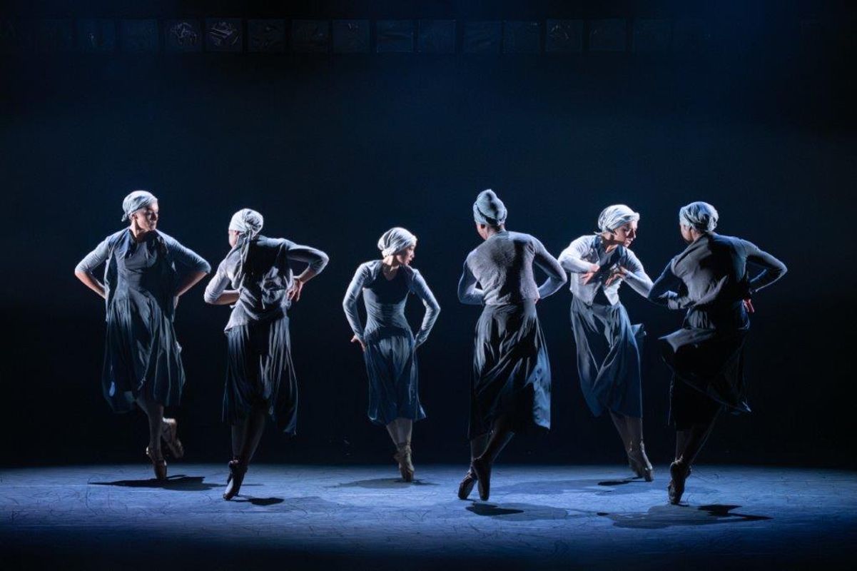 Group of dancers on stage wearing blue and grey outfits, a light casts a shadow over them