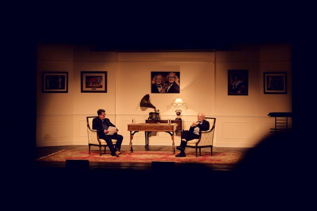 Sir David Jason on stage with another gentleman. The setting is made of two chairs, a dark wooden desk and a large rug