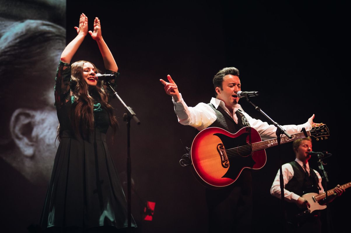 Two people stand on stage with their arms raised in a clapping motion.