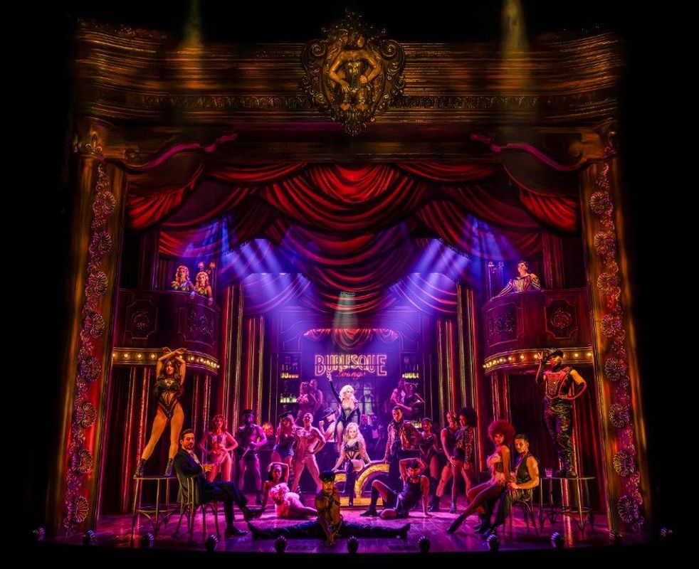 Lots of performers on stage in various different poses/positions, most are wearing corsets and fancy costumes