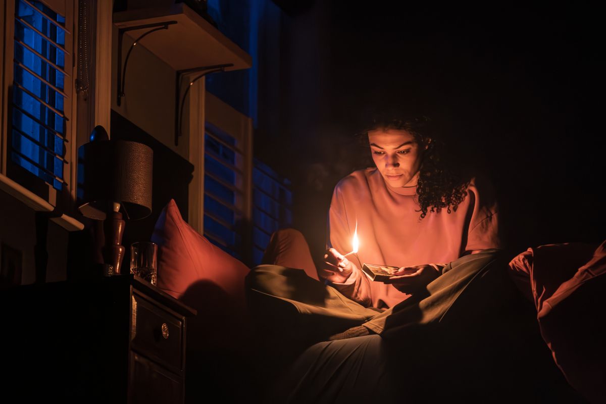 A person sits on a bed in a dimly lit room, holding a small flame, creating a warm glow on their face. The room has a calm, introspective vibe.