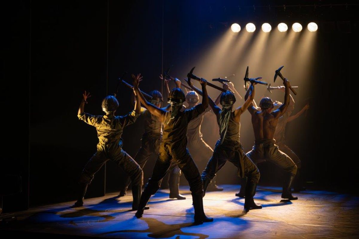 Group of dancers on stage wearing hard hats and holding pick axes above their heads