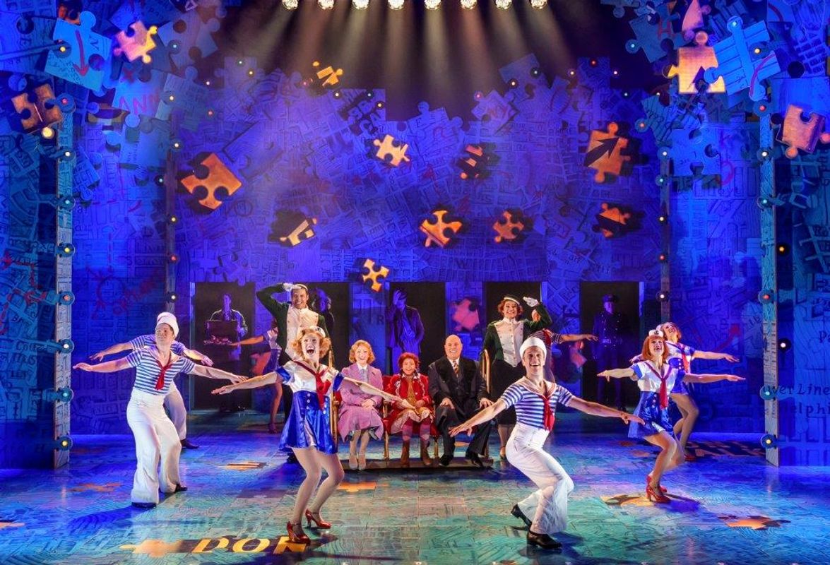 Theater scene with vibrant costumes; performers in sailor outfits dance energetically. Puzzle piece backdrop, colorful lighting creates a joyful atmosphere.