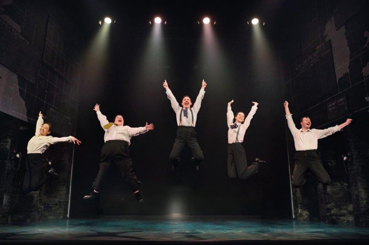 Five people jumping into the air with their arms up, all wearing white shirts, ties and black trousers