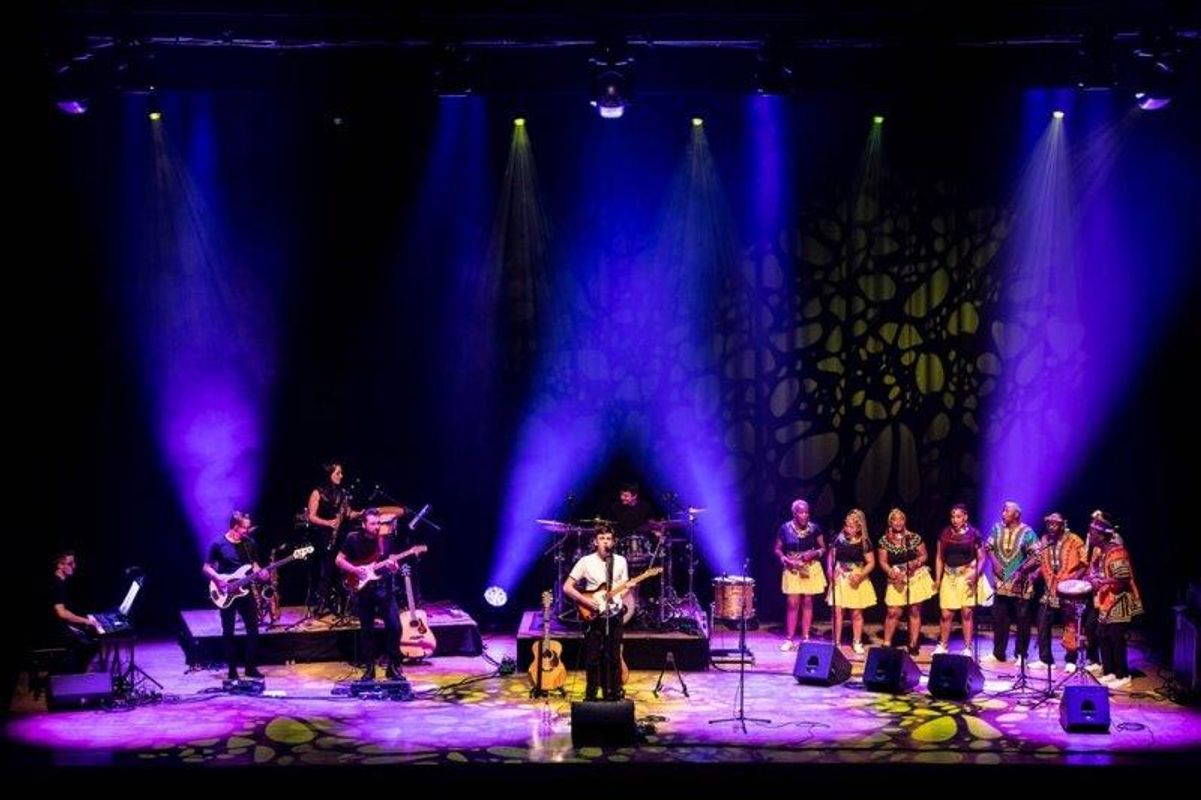 The South African Cultural Choir are singing on stage with Paul Simon. They are all wearing bright and vibrant African clothing. Paul is playing a guitar