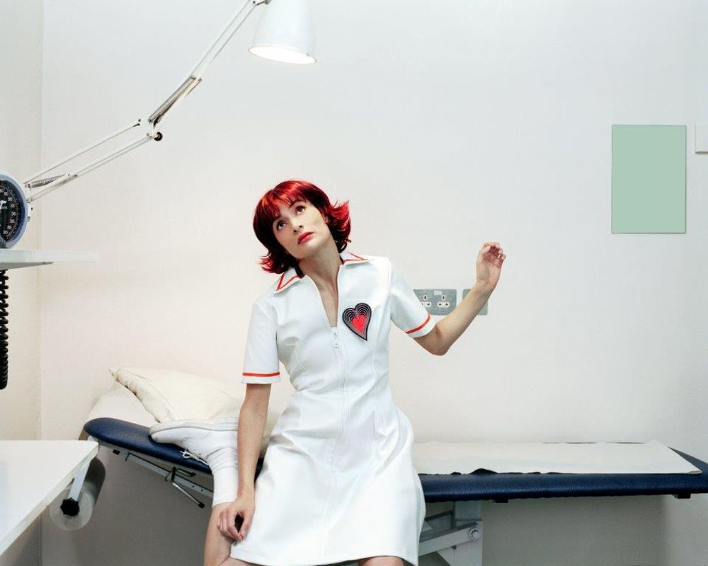 Person dressed in a white nurses outfit in a hospital room sitting on an examining bed