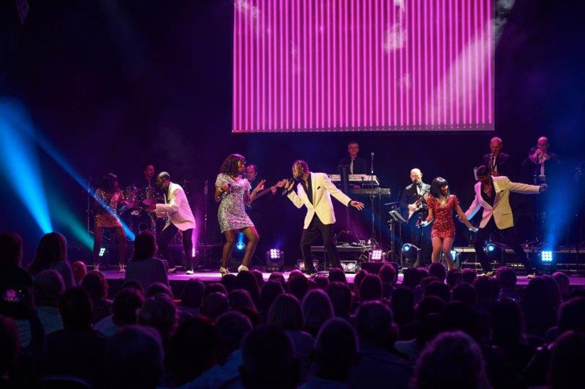 Multiple people stand on stage in brightly coloured outfits singing and dancing.