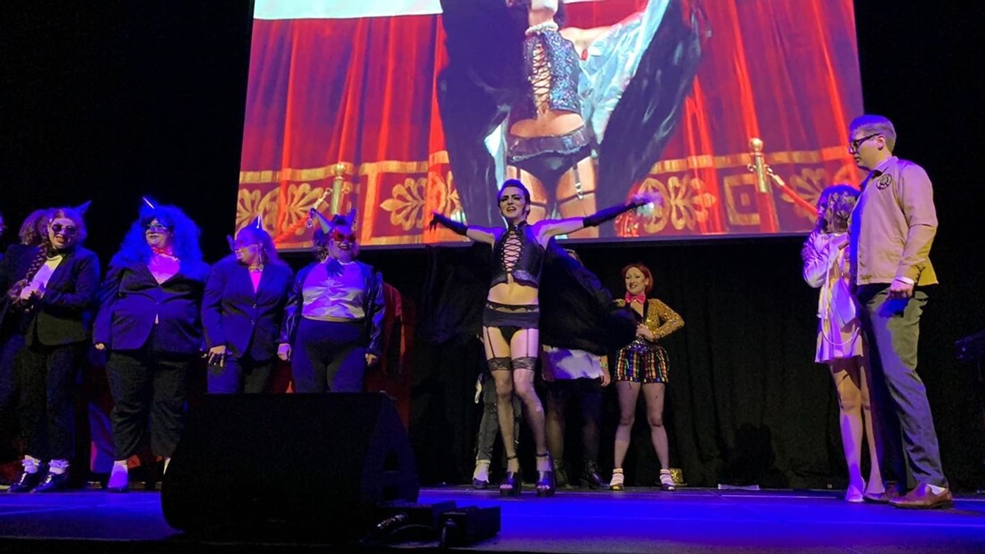 Group of people on stage. Those stood at the back are wearing black suits, white shirts and party headwear. Two people to the are looking at the people centre stage. The person in the middle has their hands up in the air, they are wearing fish net stockings, platform shoes and a corset, the people behind them are pulling a cape off the person in the middle.