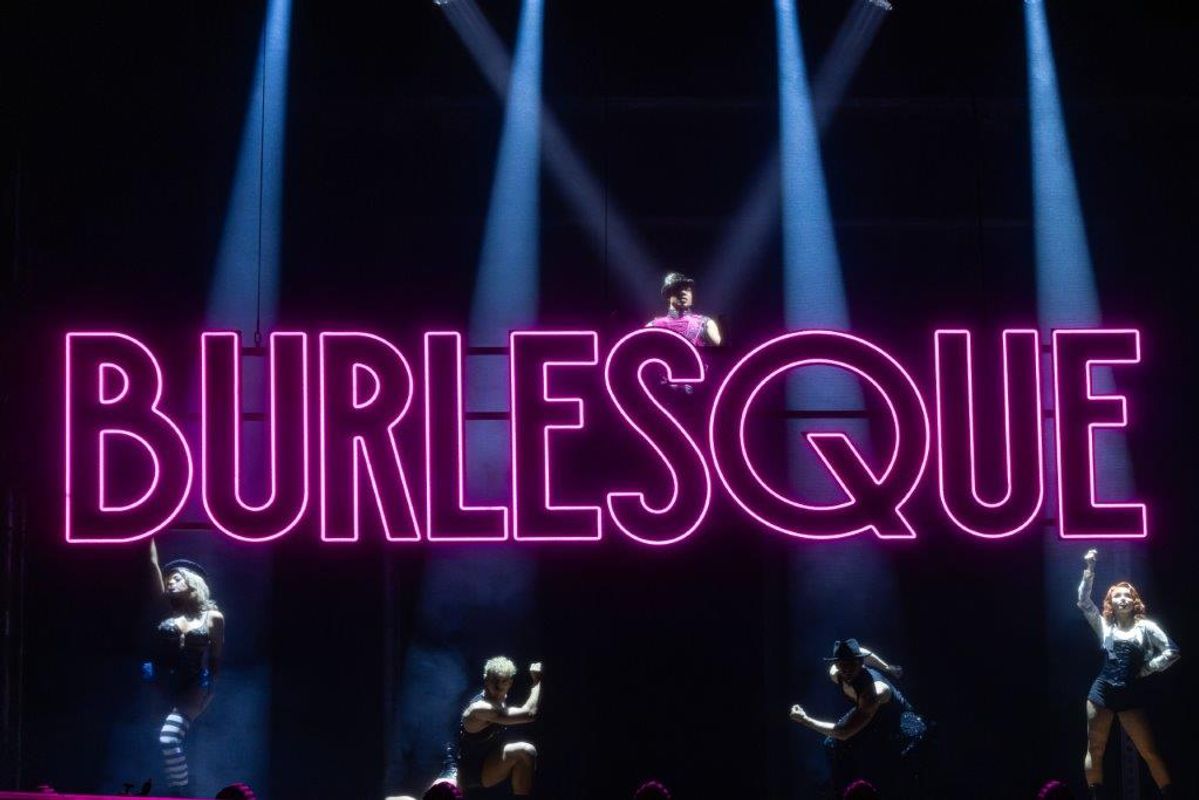 The word 'BURLESQUE' is lit up on neon lights and there are performers posing by it