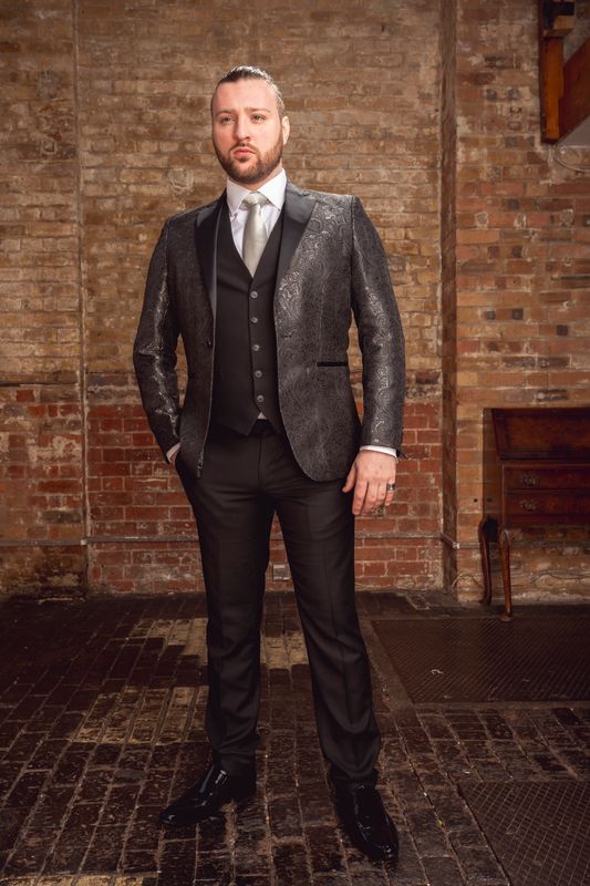 Aaron Price Lewis in a sleek, shimmering black suit and tie stands confidently in a rustic room with exposed brick walls. The atmosphere feels formal and stylish.