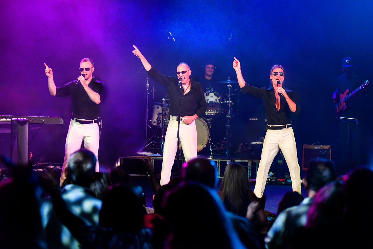 Three people dressed in white trousers, black shirts and sunglasses point their right arm in the air.
