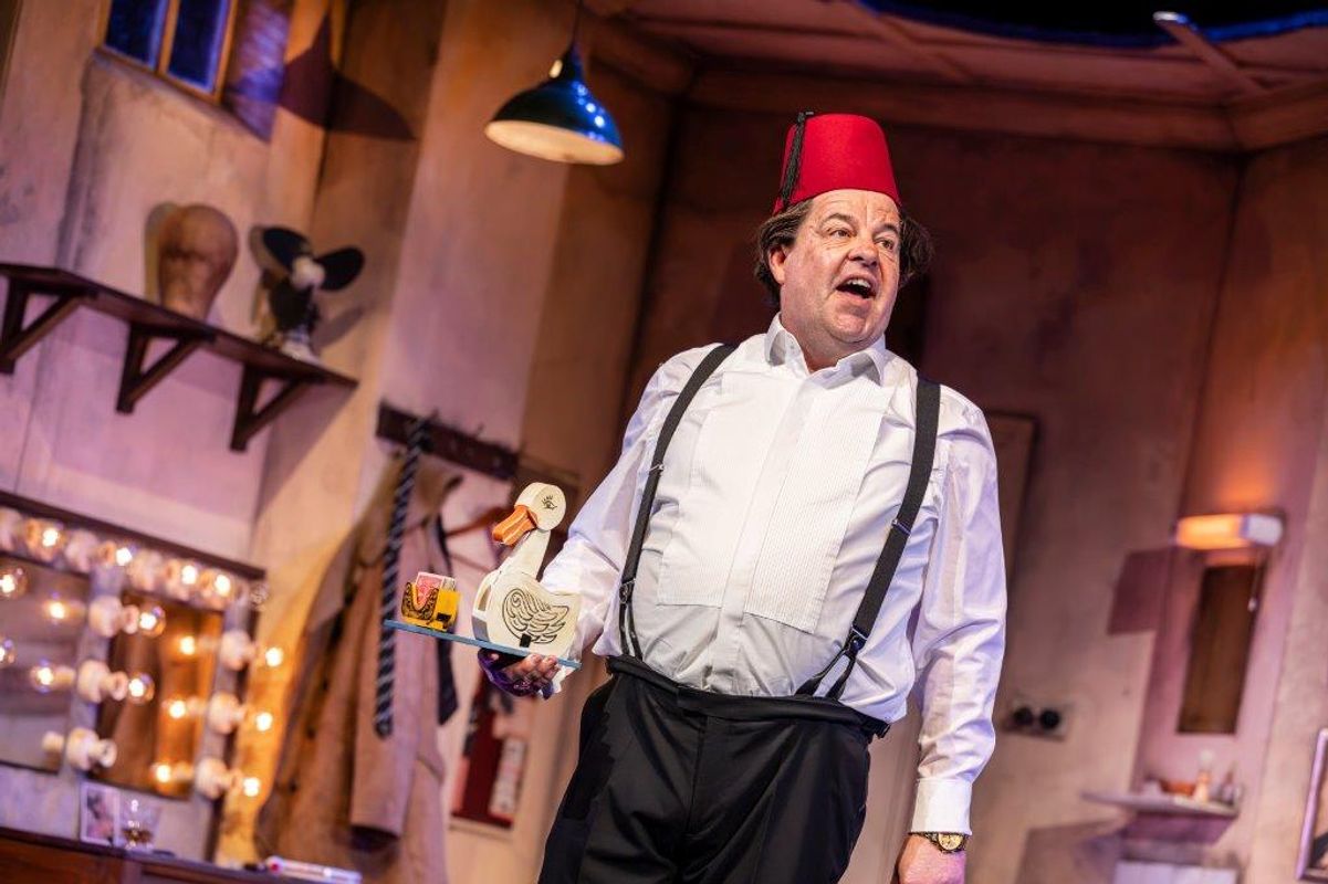 A man wearing a fez, sits onstage with an exaggerated expression. The room is cluttered and lively, featuring a vintage vanity with lights, posters, and props. The atmosphere is theatrical and whimsical, evoking humor and nostalgia. And holding a duck